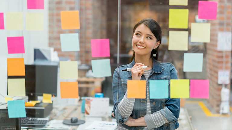 woman smiling and looking at a clear screen full of sticky notes, woman-looking-at-chalkboard-with-stuff-on-it-13-positive-affirmations-to-make-you-successful-healthista