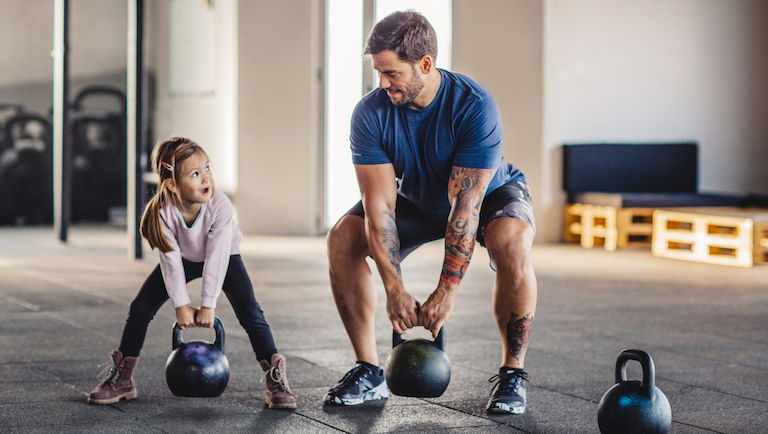 Father doing his training with kettlebells in gym while his little daughter supporting him.